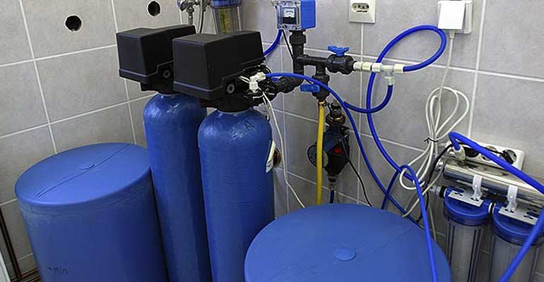 A dual tank water purification system features multiple blue tanks and cylindrical filters, connected by various pipes and hoses. The setup includes water softeners, control valves, and a digital display, all neatly arranged in a tiled room.