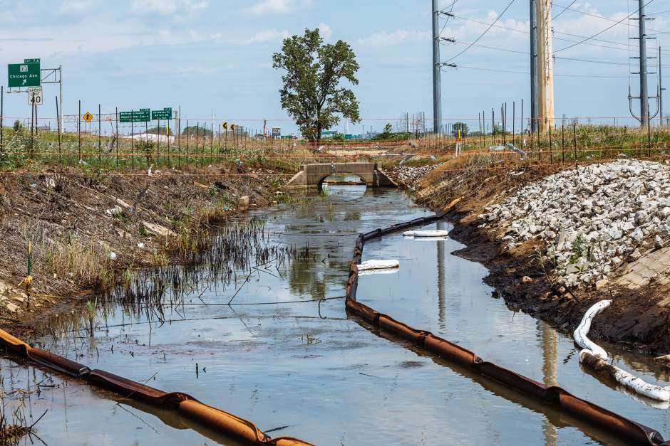 Drainage ditch with sediment barriers, surrounded by construction materials and signage, leading to a small bridge.