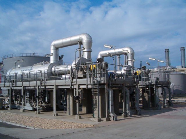 An industrial plant with large metal pipes and structures looms under a cloudy sky, equipped with Multi-Effect Distillation systems. The facility, featuring tanks, walkways, and equipment, indicates a processing or energy-related function. Gravel covers the ground around the complex.