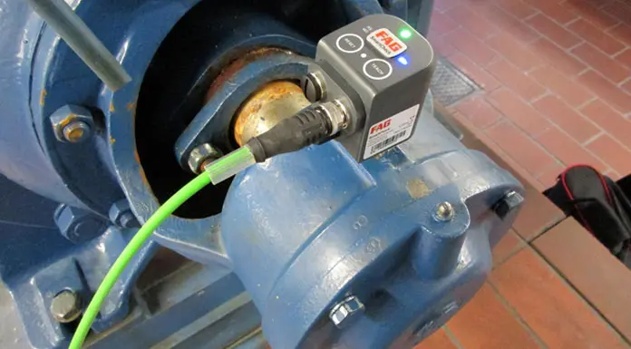 A blue industrial machine equipped with a vibration monitoring system features a sensor device connected by cable, displaying green and blue lights. Signs of wear mark the surface, while the reliability-boosting equipment stands on a tiled floor in reddish-brown.