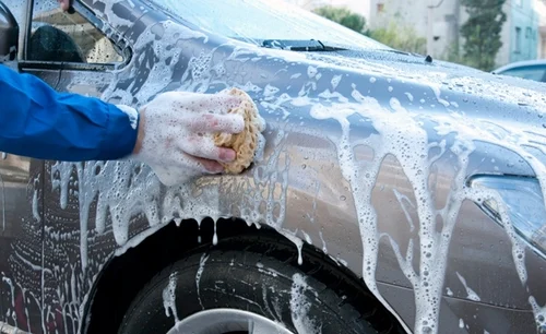 A person wearing a blue jacket and white glove uses a sponge to scrub a silver car covered in soapy foam at the car wash. The cleaning process is taking place outdoors, with blurry buildings in the background.