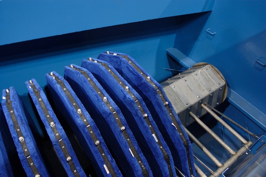 A close-up of a blue mechanical structure with multiple round, textured blue plates, reminiscent of disk filters, aligned on a metal axle. The apparatus is part of a larger machine against a blue wall, creating an industrial and technological appearance, essential for clean water systems.