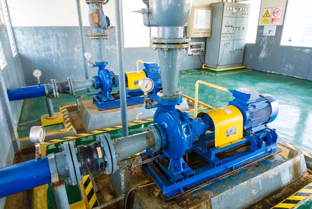 Image of an industrial setting with blue and yellow machinery, likely pumps, designed for efficient water management on a green floor. Piping connects the machinery, with gauges present. Warning signs are visible in the background on a control panel, enhancing the pump station monitoring process.