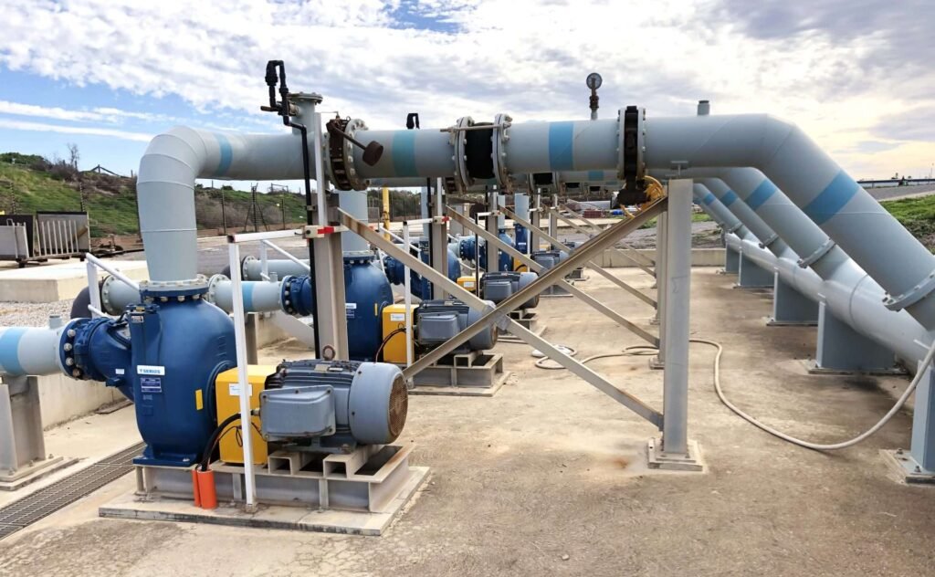 Industrial water pump station with blue machinery and large pipes under a cloudy sky.