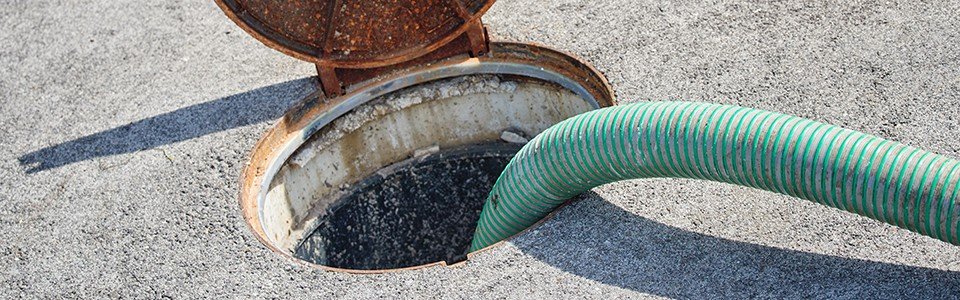 A green hose, part of an efficient solution for home plumbing, is connected to an open septic tank lid on a concrete surface for pumping or maintenance work. The rusty lid is partially open, revealing the circular opening of the tank.