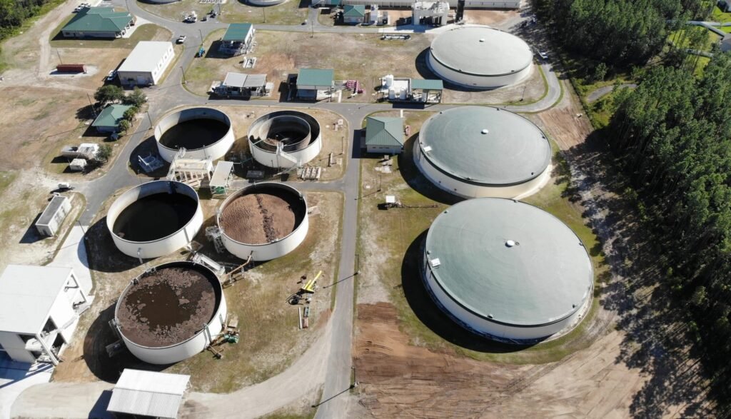 Aerial view of a wastewater treatment plant with large circular tanks and surrounding buildings amidst greenery.