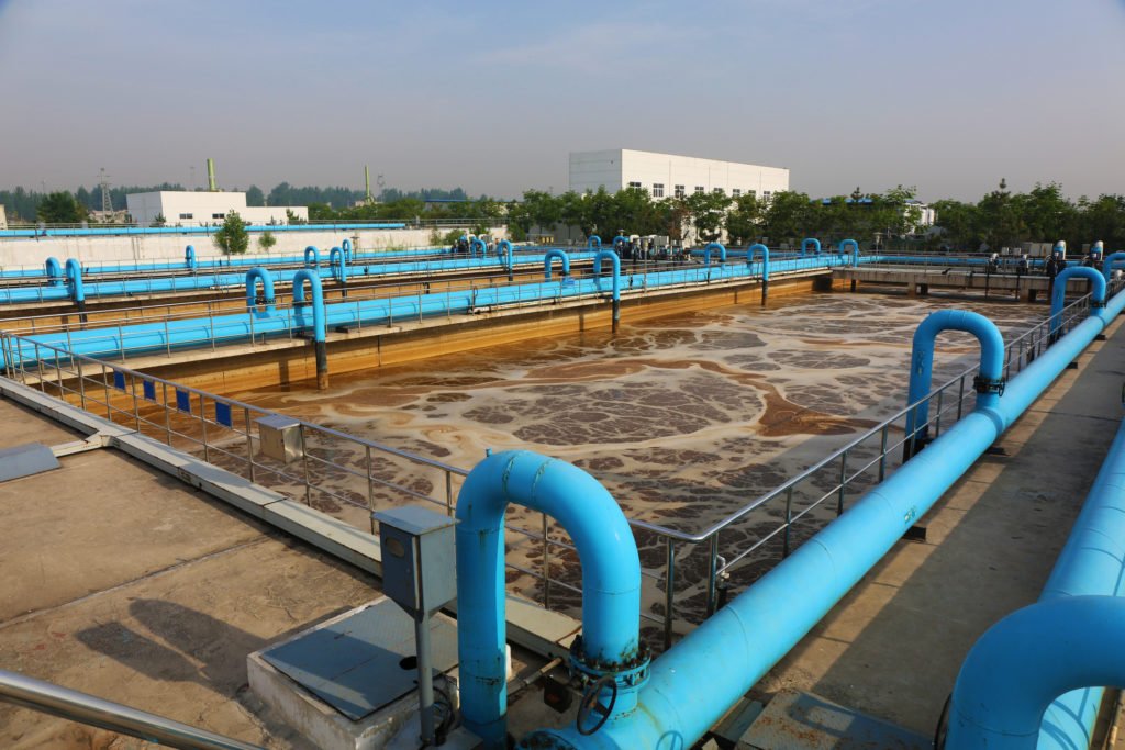 Wastewater treatment facility with large basins and blue pipes under a clear sky.