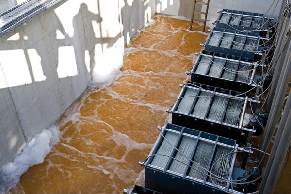 At a wastewater treatment facility, large rectangular tanks with reddish-brown water showcase advanced purification technologies. Cables and equipment surround the foamy, agitating water, while shadows of people and a ladder dance on the concrete wall.