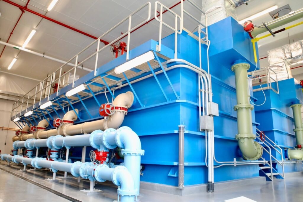 Large industrial blue filtration tanks, crucial for clean operations, are connected by a network of pipes and valves in a well-lit facility. Metal walkways above ensure efficient maintenance. Overhead lights and red pipes highlight the robust infrastructure of these water purification systems.