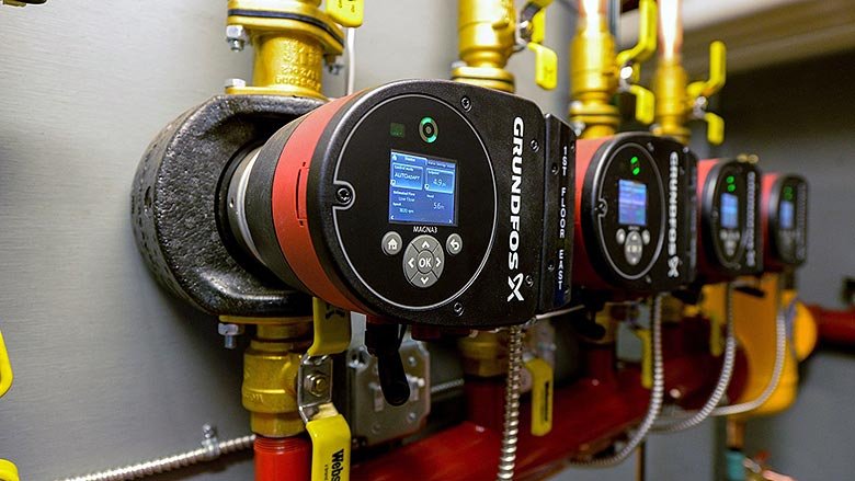 A row of black and red Grundfos circulator pumps, attached to yellow pipes, are mounted on a wall. Each pump displays a digital screen with operational information. This efficient solution is part of an industrial or commercial plumbing system.