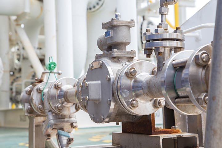 Close-up of industrial metal piping with valves and flanges, possibly part of a gas or liquid transport system. The silver-colored pipes, essential for positive displacement pumps, are interconnected and have various fittings, mounted on a platform with some visible rust.