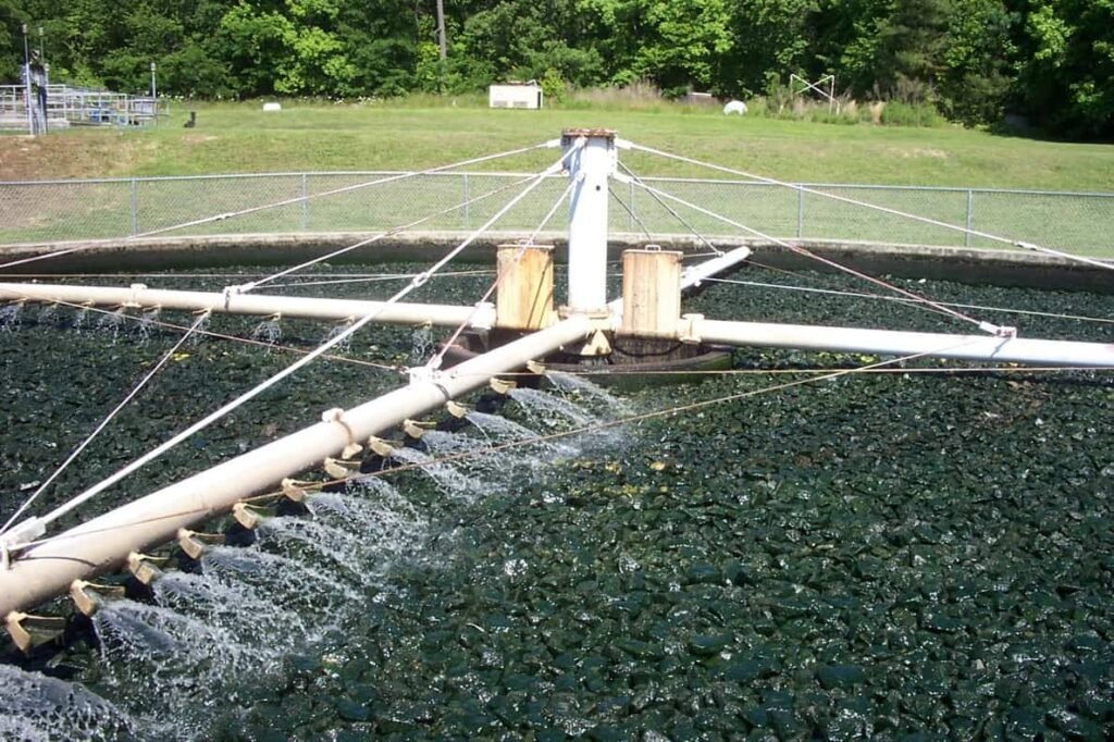 A circular trickling filter with water being sprayed over a bed of rocks showcases successful restoration. It features a central pivot and radiating arms distributing water, surrounded by a fence and trees in the background.